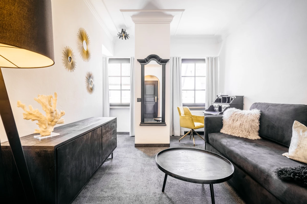 Modernes, helles Hotelzimmer mit grauem Sofa, gelbem Sessel und dekorativen Wandelementen. Weiße Wände, große Fenster und ein runder Couchtisch prägen den minimalistischen, einladenden Wohnbereich. Eine stehende Lampe und eine Kommode vervollständigen das zeitgenössische Interieur.