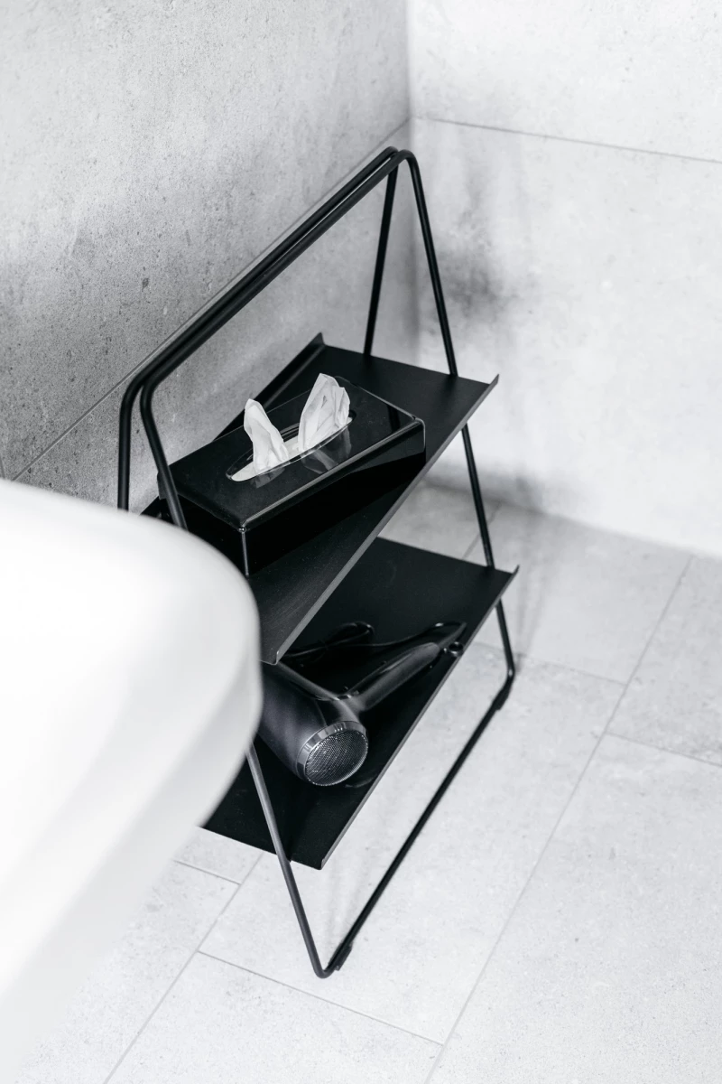Modernes, minimalistisches schwarzes Regal mit zwei Ebenen, auf dem eine weiße Tissuebox und ein schwarzer Ball liegen. Das Regal steht in einer monochromen, hellen Ecke eines Raumes.