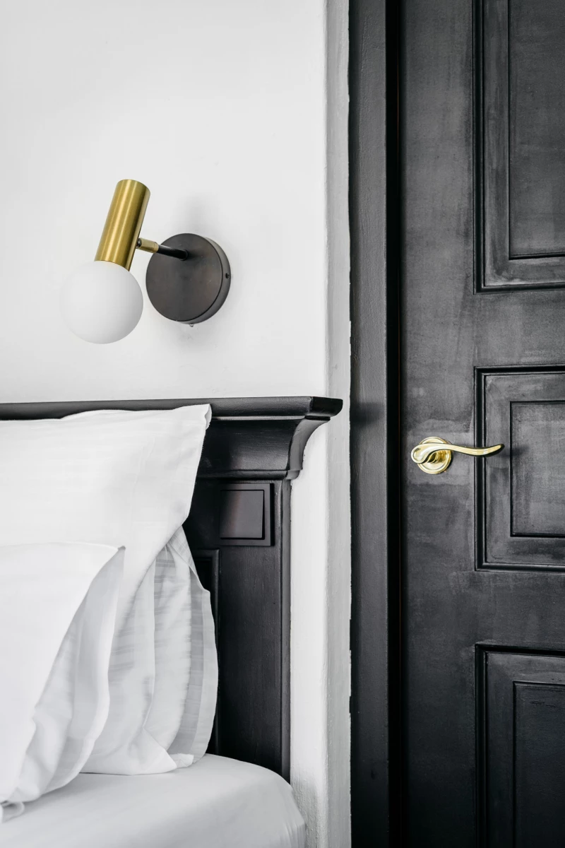 Modernes Hotelzimmer mit elegantem Schlafbereich, schwarzer Tür mit goldener Klinke und einer goldenen Wandleuchte an der weißen Wand, die minimalistisches und stilvolles Interieurdesign betont.